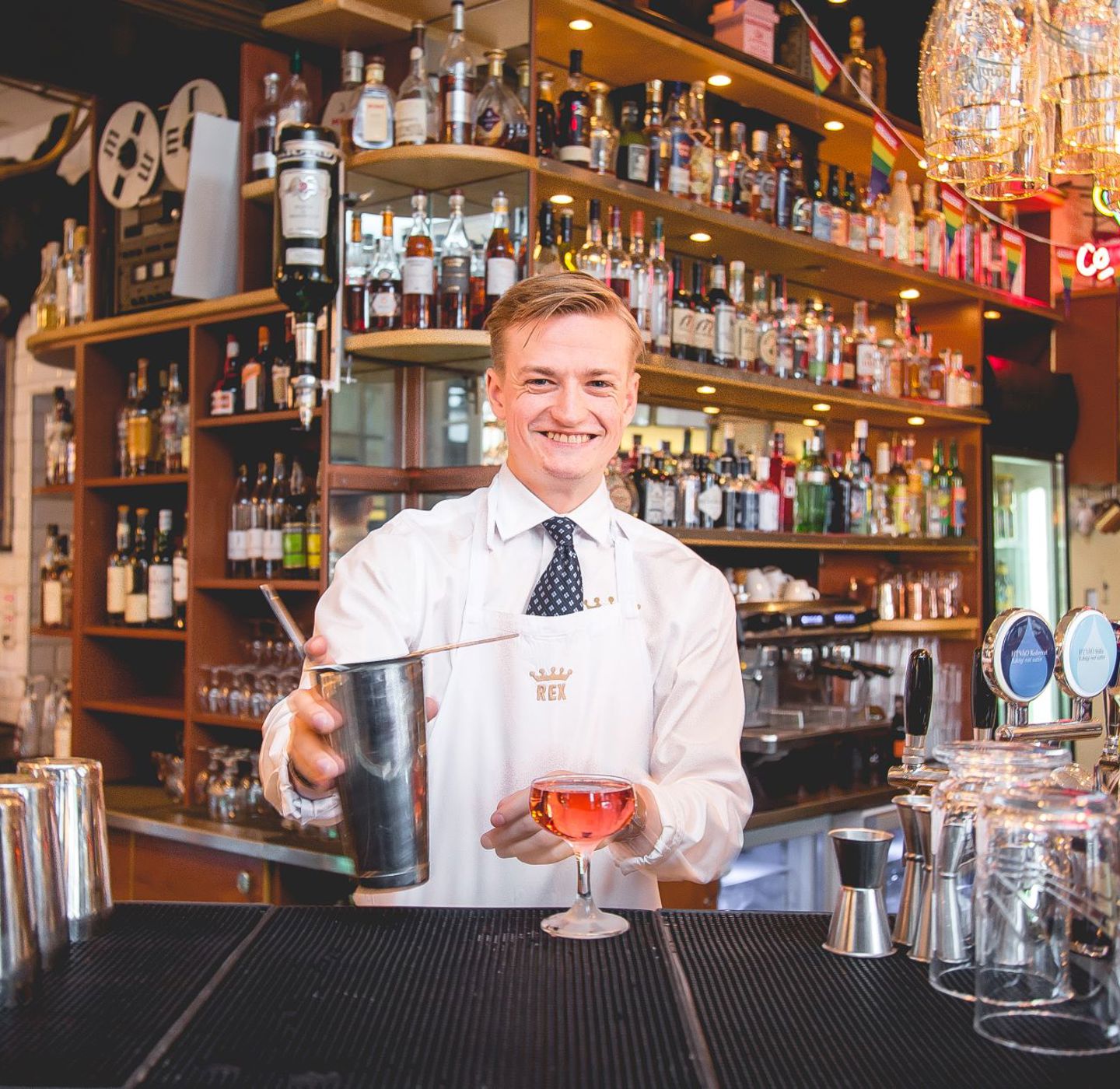 En glad man med vit klädsel och slips står i en bar och häller upp en drink. 