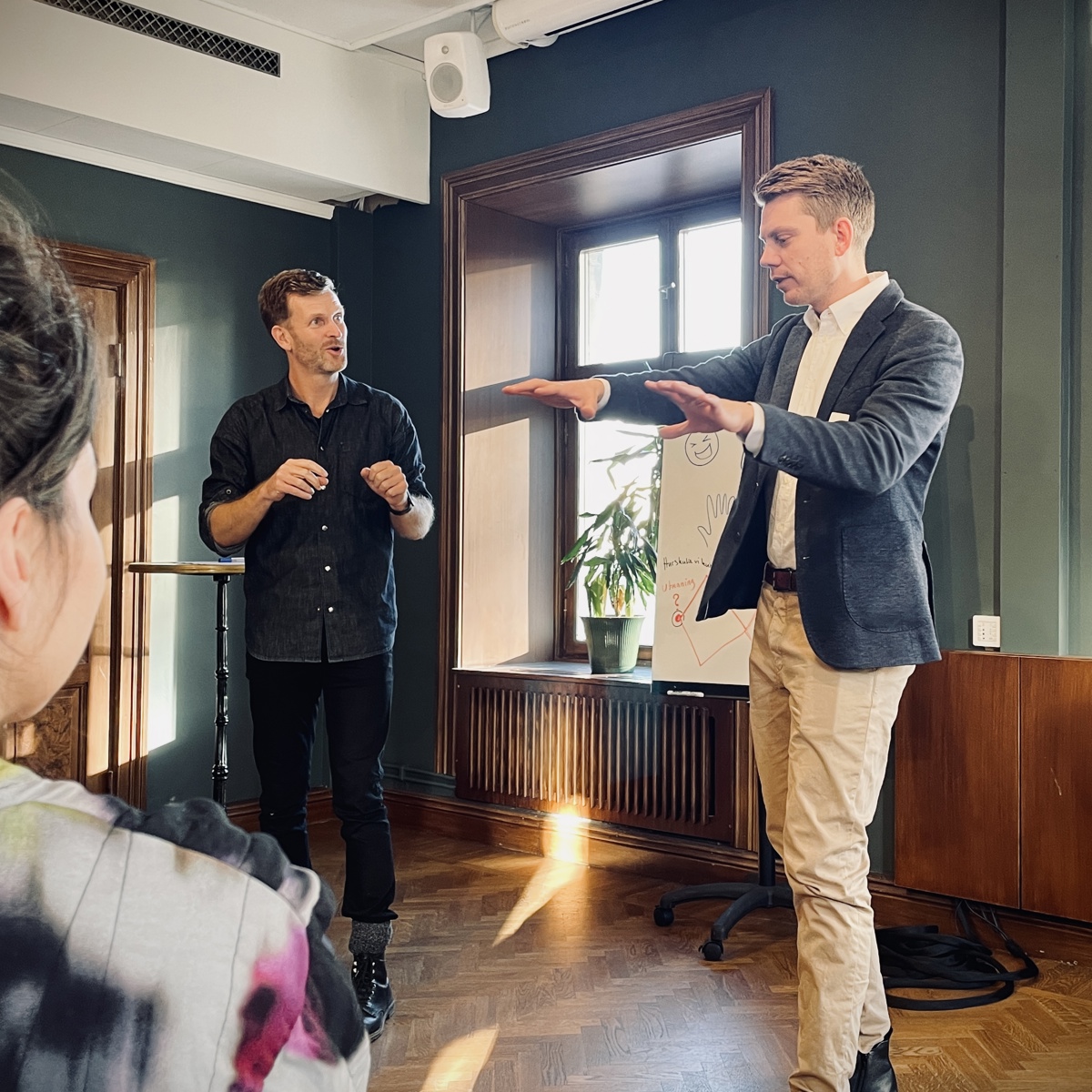Två män står framför en samling och gestikulerar, en kvinnas huvud i förgrunden. 