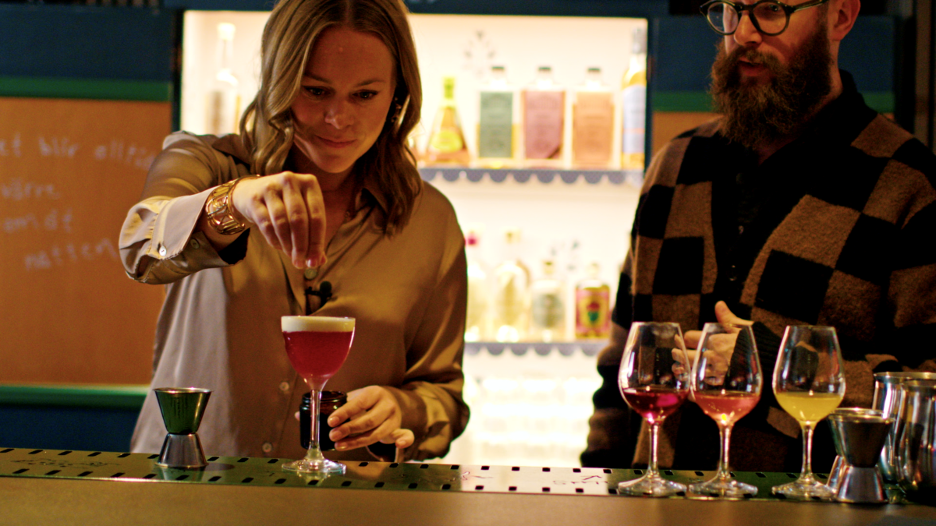 Maya förbereder en drink på Facit bar i tv-serien Drinking With Joe & Maya. Foto: Lycky Day Pictures En kvinna förbereder en drink och en man tittar på.
